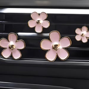 Può includere: Quattro decorazioni per bocchette d'aria per auto a forma di fiore rosa, con bordi e centri dorati. I fiori sono disposti su una bocchetta nera, creando un accessorio decorativo ed elegante.