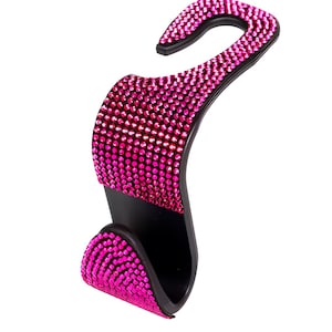 May include: A black car seat headrest hook adorned with vibrant pink rhinestones. The hook is designed in a curved, S-shape, with the pink rhinestones covering the front-facing surfaces. The hook is designed to hang items in a car.