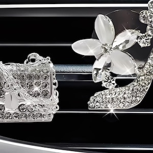 May include: Two car air fresheners: a silver handbag with a chain strap and a high-heeled shoe with a flower accent. Both are encrusted with sparkling crystals, set against a black background.