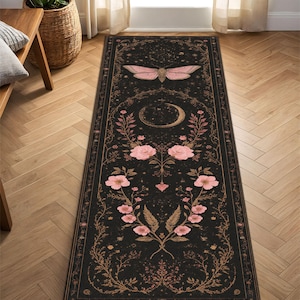 May include: Black rectangular rug with a detailed floral and celestial design. A pink moth is at the top, with a crescent moon and flowers below. The rug has a dark background with a decorative border.