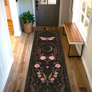 May include: Black runner rug with a celestial design. The rug features a large pink moth at the top, a crescent moon, and floral and botanical elements in pink and gold. The rug is rectangular and placed in a hallway.