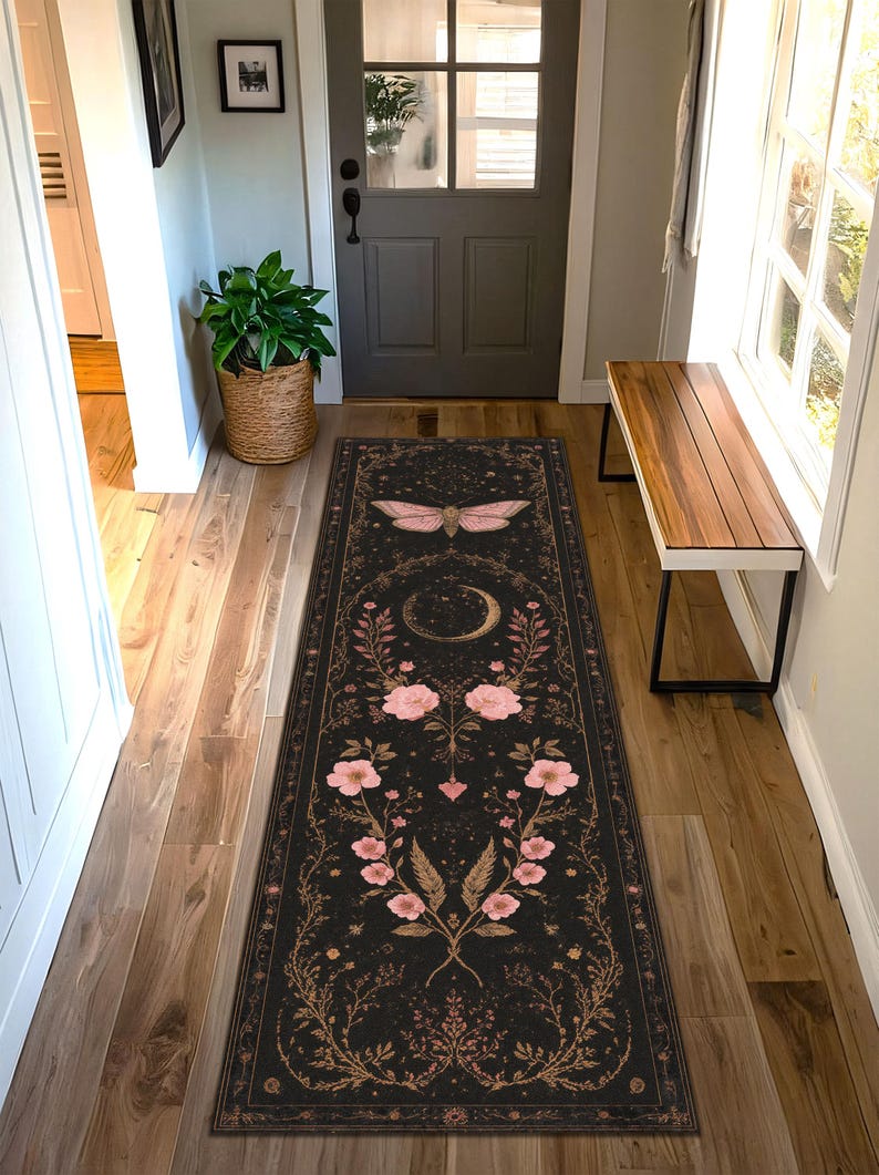 May include: Black runner rug with a celestial design. The rug features a large pink moth at the top, a crescent moon, and floral and botanical elements in pink and gold. The rug is rectangular and placed in a hallway.