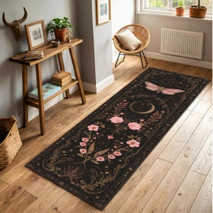 May include: A long, rectangular black rug with a floral and celestial design. Pink flowers and a moth are featured on the rug, along with a crescent moon. The rug is placed on a wooden floor.