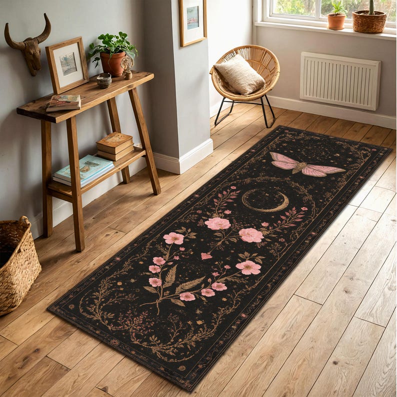 May include: A long, rectangular black rug with a floral and celestial design. Pink flowers and a moth are featured on the rug, along with a crescent moon. The rug is placed on a wooden floor.
