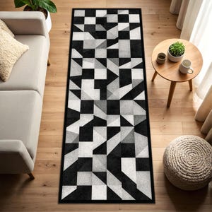 May include: A rectangular rug with a black and white geometric design. The rug has a repeating pattern of squares, triangles, and other shapes in shades of black, white, and grey. It is placed on a wooden floor.