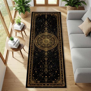 May include: Black rectangular rug with a gold celestial design. The rug features a central compass rose design, surrounded by stars, moons, and geometric patterns. The rug is placed on a hardwood floor, with a sofa and side tables in the background.