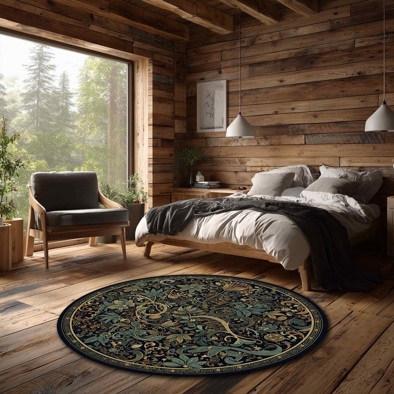 Op de afbeelding: Een rond vloerkleed met een donkere achtergrond en een gedetailleerd bloemenpatroon in groen, goud en blauw. Het kleed ligt op een houten vloer in een slaapkamer, met een bed, stoel en groot raam.