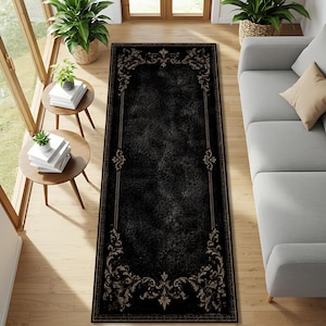 May include: A rectangular runner rug featuring a deep black centre with a distressed, textured appearance. The rug is framed by a decorative border in muted beige, showcasing intricate scrolling floral and leaf patterns at each corner.