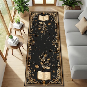 May include: A rectangular black rug with a gold floral and book design. The rug features an open book at the top and bottom, with a central floral motif and decorative border. The rug is placed on a wooden floor.