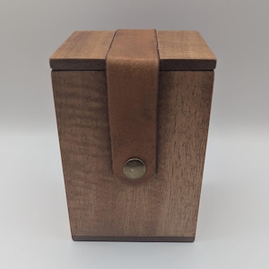 May include: A wooden box with a hinged lid and a leather strap closure. The box is made of dark-stained wood and has a simple, minimalist design. The brown leather strap is secured with a brass-coloured stud.