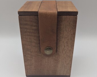 Könnte beinhalten: Eine Holzbox mit Klappdeckel und Lederriemenverschluss. Die Box ist aus dunkel gebeiztem Holz gefertigt und hat ein schlichtes, minimalistisches Design. Der braune Lederriemen ist mit einem messingfarbenen Knopf befestigt.
