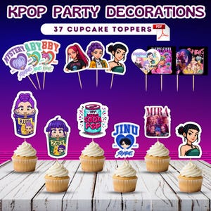 May include: K-Pop party decorations featuring 37 cupcake toppers. The toppers showcase various K-Pop group illustrations and text, including "MYSTERY", "ABYBBY", "RUMI", "ZOEY", and "JINU". Cupcakes with white frosting are arranged on a wooden surface.