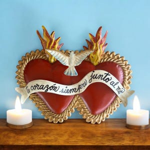 Może przedstawiać: Czerwona dekoracja ścienna w kształcie serca ze złotą obwódką, płomieniami i gołębiem. Na banerze nad sercami widnieje napis "Tu corazón siempre junto al mío". Dwie białe świece na drewnianej powierzchni.