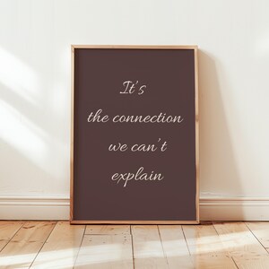 Può includere: Una stampa incorniciata con la scritta "It's the connection we can't explain" in bianco. Lo sfondo è marrone scuro e la cornice è in legno chiaro. La stampa è appoggiata a una parete bianca su un pavimento in legno.