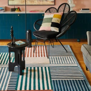 May include: A modern living room featuring a black chair with a patterned pillow, a striped rug in shades of blue, green, and red, and a small black side table. A painting hangs on the wall above a blue cabinet.