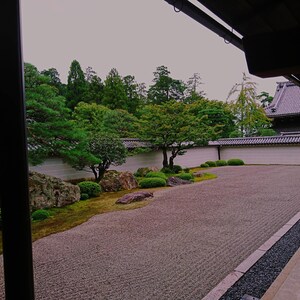 以下が含まれることがあります： 砂利の中庭、手入れの行き届いた緑、瓦屋根の伝統的な建物が特徴の、穏やかな日本の庭園の風景。 構図は暗い木造構造で構成され、奥行きと静けさを生み出しています。 色使いは落ち着いており、緑、茶色、灰色が使われています。