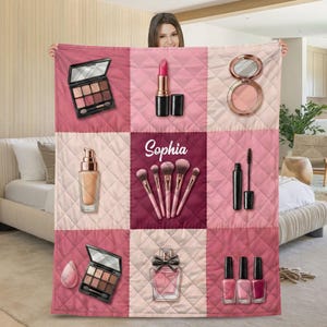 Op de afbeelding: Een gewatteerde deken met een raster van make-up items in roze en bordeaux tinten. Het ontwerp omvat oogschaduwpaletten, lippenstift, foundation, make-up borstels, mascara, blush, parfum, nagellak en een make-up spons. De naam "Sophia" staat erop.