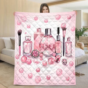 Op de afbeelding: Een roze gewatteerde deken met een ontwerp van parfumflesjes, make-up borstels en roze edelstenen. Het ontwerp bevat een grote parfumfles met een strik, een ring en roze bollen. De achtergrond is wit met een ruitpatroon.