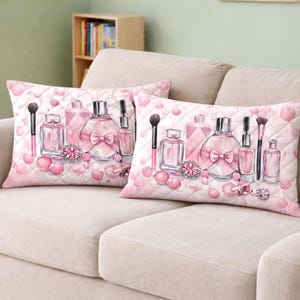 Op de afbeelding: Twee roze decoratieve kussens met een parfum- en make-upontwerp. De kussens hebben illustraties van parfumflesjes, make-upborstels en roze juwelen op een gewatteerde achtergrond. De kussens zijn rechthoekig en liggen op een beige bank.
