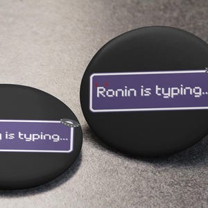 Peut inclure: Deux boutons ronds noirs avec le texte "goreboy is typing..." et "Ronin is typing..." en blanc sur fond violet. Les boutons ont une finition brillante et un petit détail argenté.