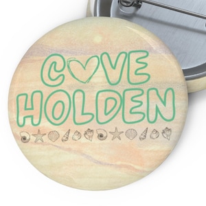 May include: A round button with the words "COVE HOLDEN" in green, with a heart replacing the "O" in "COVE". The background has a sandy beach design with small seashell and starfish illustrations.