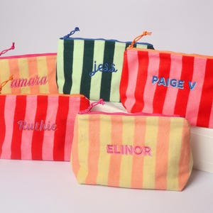 May include: Five rectangular zippered pouches featuring vertical stripes in various color combinations including red, pink, orange, yellow, and green. Each pouch is embroidered with a name: amara, jess, PAIGE V, Ruthie, and ELINOR.