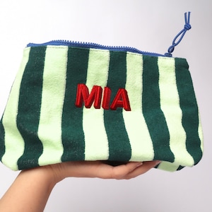 May include: A zippered pouch featuring vertical stripes in dark green and light green. The name MIA is embroidered in red thread on the front. The pouch has a blue zipper closure with a matching blue cord pull attached to the zipper slider.