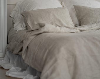 Puede incluir: Una cama con ropa de cama de lino en tonos neutros. La cama tiene una funda nórdica y fundas de almohada de color beige claro, con fundas de almohada blancas con volantes y una falda de cama blanca. La escena está bañada en luz suave y natural.