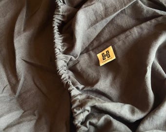 May include: Dark gray fitted sheet with elastic edges. A small, rectangular label with a logo is sewn onto the sheet. The sheet is made of a soft, textured fabric, likely linen or cotton. The overall theme is bedding.