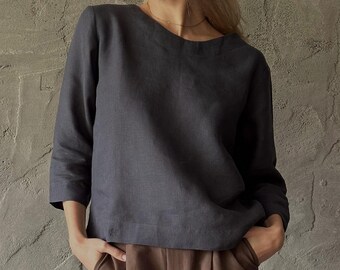 Puede incluir: Una blusa de lino gris oscuro con mangas tres cuartos y cuello redondo. La blusa se combina con pantalones marrones. La modelo lleva un collar dorado. La tela parece suave y fluida, lo que sugiere un ajuste cómodo.