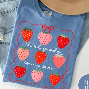 May include: A light blue t-shirt with a strawberry design and the text "third grade is my jam." The shirt is paired with light wash blue jeans and a tan hat. A silver necklace is visible.