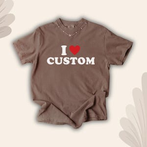 May include: A brown t-shirt with the text "I &hearts; CUSTOM" in white, with a red heart replacing the word "love". The shirt has short sleeves and a crew neck. The shirt is laid flat on a neutral surface.