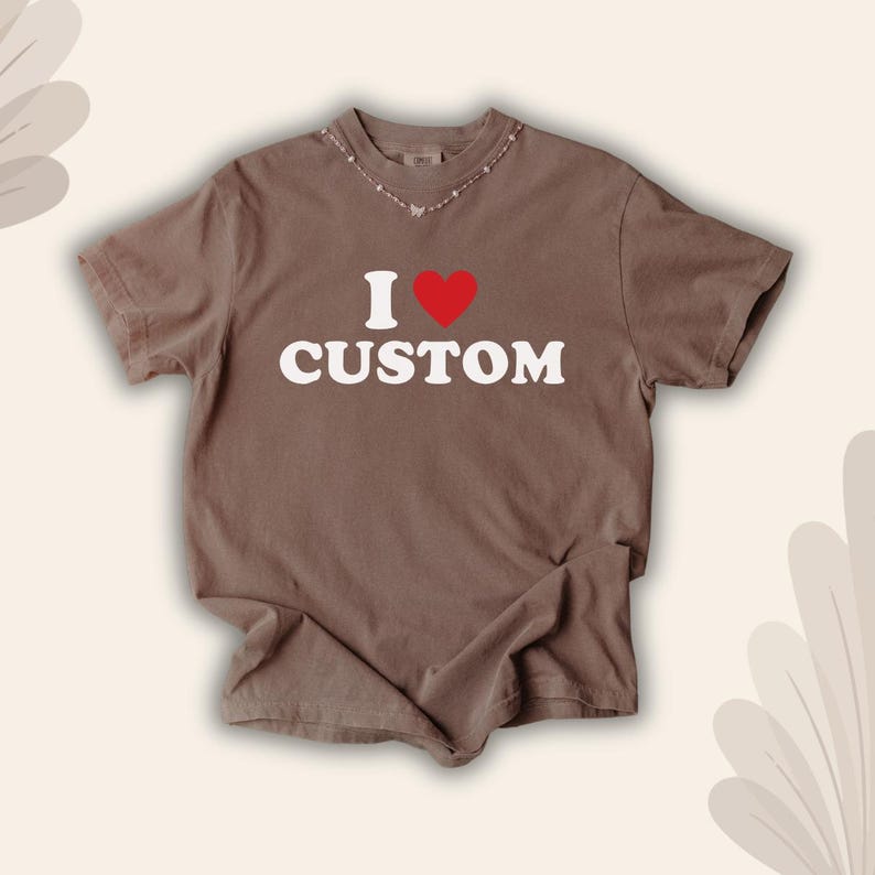 May include: A brown t-shirt with the text "I &hearts; CUSTOM" in white, with a red heart replacing the word "love". The shirt has short sleeves and a crew neck. The shirt is laid flat on a neutral surface.