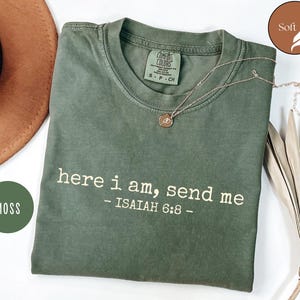 Könnte beinhalten: Ein moosgrünes T-Shirt mit dem weißen Text "here i am, send me - ISAIAH 6:8". Das Shirt hat einen Rundhalsausschnitt und kurze Ärmel. Eine goldene Halskette mit Anhänger liegt auf dem Shirt. Ein brauner Hut ist im Hintergrund zu sehen.