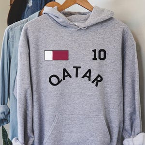 K&ouml;nnte beinhalten: Ein grauer Kapuzenpullover mit dem Wort "QATAR" in schwarzem, gebogenem Text. &Uuml;ber dem Text befindet sich ein weinrotes und wei&szlig;es Rechteck und die Zahl "10" in Schwarz. Der Kapuzenpullover h&auml;ngt an einem Holzb&uuml;gel.