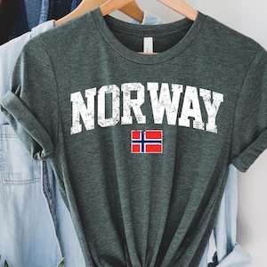 以下が含まれることがあります： 「NORWAY」の文字が白でかすれたようにプリントされた、濃い緑色のTシャツ。小さなノルウェーの国旗のグラフィックもあしらわれています。他の衣類と一緒に木製のハンガーに掛けられています。