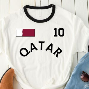 K&ouml;nnte beinhalten: Wei&szlig;es T-Shirt mit schwarzem Besatz und dem Wort "QATAR" in gro&szlig;en schwarzen Buchstaben. Die Nummer 10 und eine Katar-Flagge sind ebenfalls aufgedruckt. Hellblaue Jeans, braune Stiefel und Sonnenbrille sind ebenfalls im Bild.