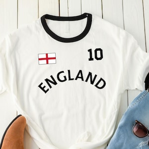 May include: A white ringer t-shirt with black trim, featuring the text ENGLAND, the number 10, and the England flag. The shirt is paired with blue denim jeans and a pair of brown suede ankle boots with dark brown soles and dark brown sunglasses.