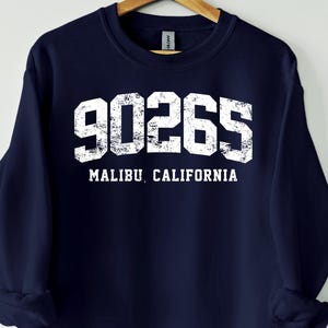 May include: A navy blue sweatshirt displaying the white text "90265 MALIBU, CALIFORNIA". The sweatshirt is suspended from a wooden hanger. The text has a distressed, vintage appearance.