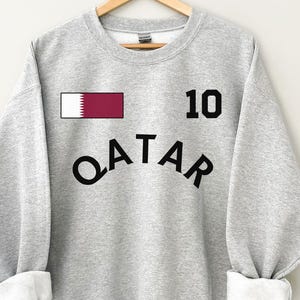 May include: A light gray sweatshirt with the word "QATAR" in black arched lettering. Above the lettering is the number "10" in black, and a small rectangle with the Qatar flag. The sleeves are cuffed with white fabric.