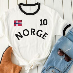 May include: White t-shirt with black trim, the word "NORGE", the number 10, and the Norwegian flag. The shirt is paired with blue jeans, brown boots, and sunglasses.