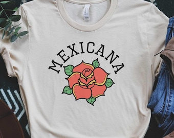 May include: Cream-colored t-shirt with the word "Mexicana" above a red rose graphic. The shirt is styled with blue jeans, brown boots, and a brown hat. The design is a casual, bohemian style.