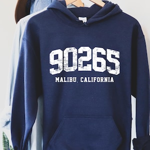 May include: A navy blue hooded sweatshirt featuring the white text "90265 MALIBU CALIFORNIA". The garment has a front pocket and a drawstring hood. A brown hat and denim jacket are visible in the background.
