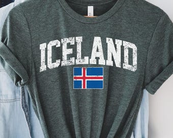Puede incluir: Una camiseta verde oscuro con la palabra "ICELAND" en letras blancas desgastadas y la bandera islandesa. La camiseta está colgada en una percha de madera con otras prendas.