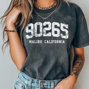 May include: Dark gray t-shirt with the text "90265 MALIBU CALIFORNIA" in white block letters. The shirt has a vintage, worn look and is paired with light blue denim jeans.