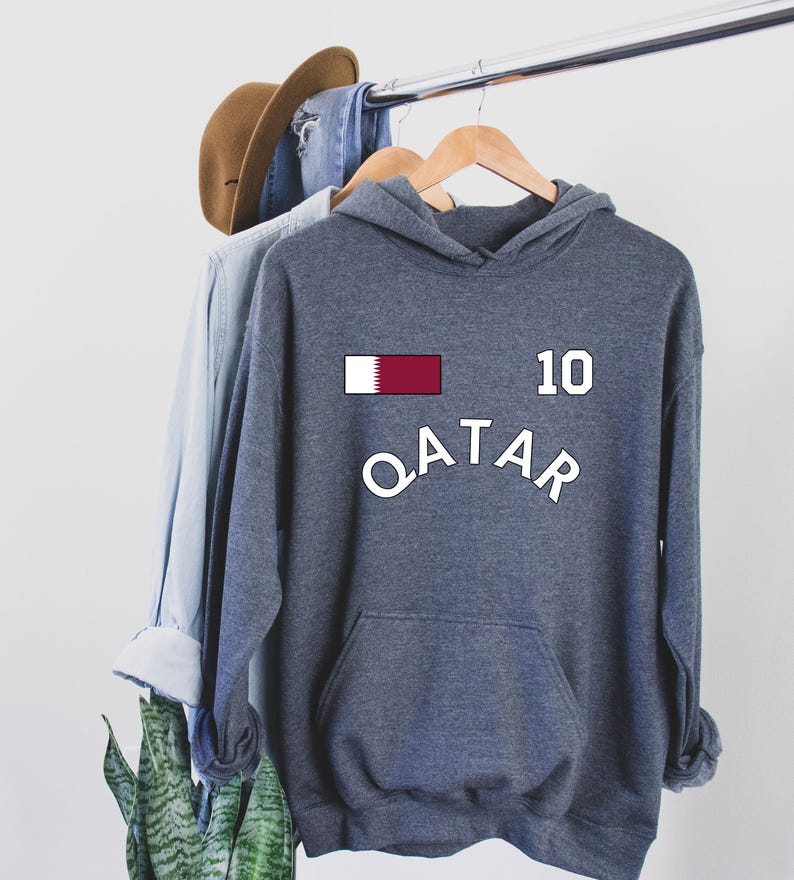 Pu&ograve; includere: Una felpa con cappuccio grigia con tasca a marsupio, con la scritta "QATAR" in testo bianco arcuato. Sopra il testo c'&egrave; il numero "10" e un disegno a bandiera rettangolare con strisce bianche e marroni. La felpa con cappuccio &egrave; appesa a una gruccia di legno.