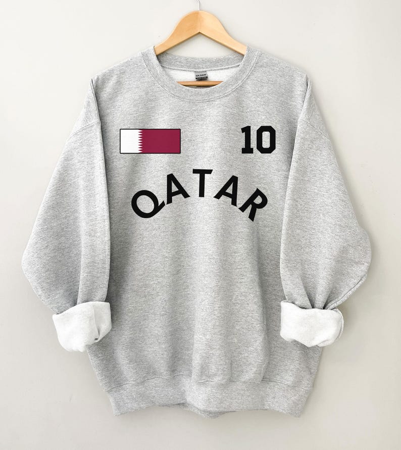May include: A light gray sweatshirt with the word "QATAR" in black arched lettering. Above the lettering is the number "10" in black, and a small rectangle with the Qatar flag. The sleeves are cuffed with white fabric.