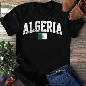 以下が含まれることがあります： 「ALGERIA」の文字が白くかすれた黒いTシャツと、小さなアルジェリアの国旗。木製の表面に、茶色の靴とブルージーンズが添えられています。