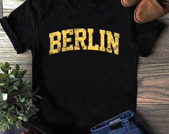 May include: Black t-shirt with the word "BERLIN" printed in distressed yellow lettering. The shirt is displayed on a wooden surface, with brown shoes and blue jeans visible in the background. A small plant is in the lower left corner.