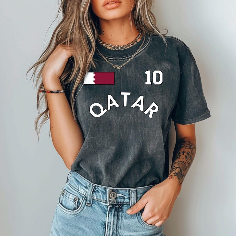 Puede incluir: Camiseta gris oscuro con la palabra "QATAR" en letras blancas. El n&uacute;mero "10" est&aacute; encima de la palabra, y una bandera de Qatar est&aacute; en el pecho izquierdo. La camiseta se lleva con vaqueros azules.